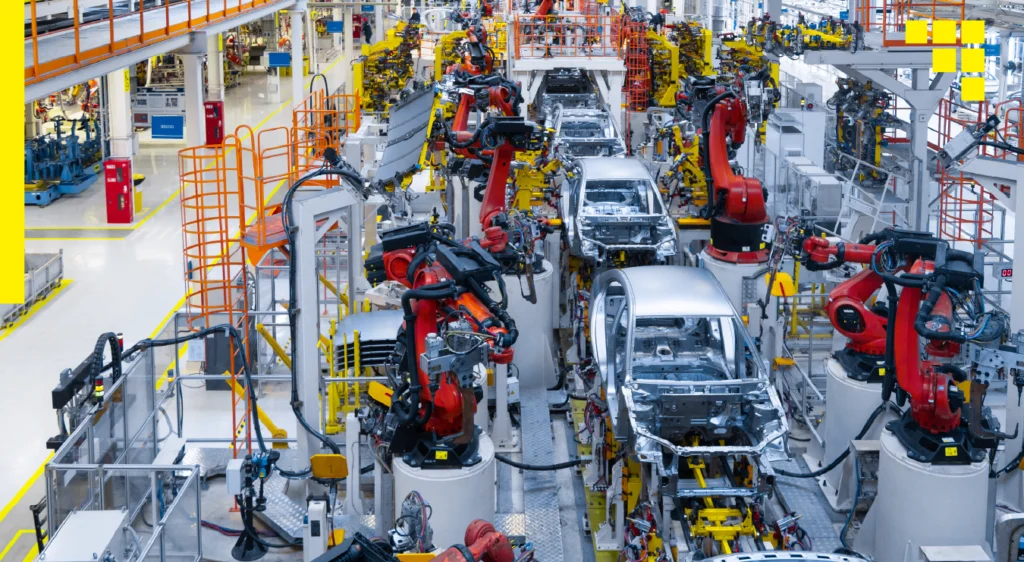 A busy automotive assembly line with multiple red robotic arms working on the chassis of cars, surrounded by various industrial equipment and machinery.